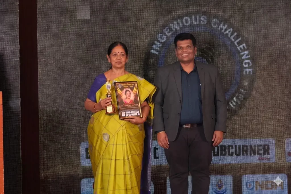Mrs. Lakshmi Ramachandran and Mr. Sivakumar, LIC agents from Chennai, standing together on stage at an award ceremony. Mrs. Lakshmi is wearing a yellow saree and proudly holding a golden trophy and a framed award certificate, while Mr. Sivakumar stands beside her smiling in a grey blazer and dark blue shirt. The background features a dark digital display screen.